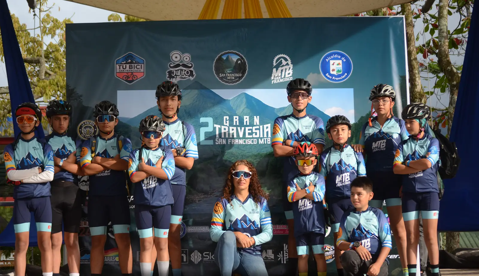 Niñas, niños y jóvenes de la escuela de ciclomontañismo con casco y uniforme, frente al telón de la Gran Travesía San Francisco MTB.